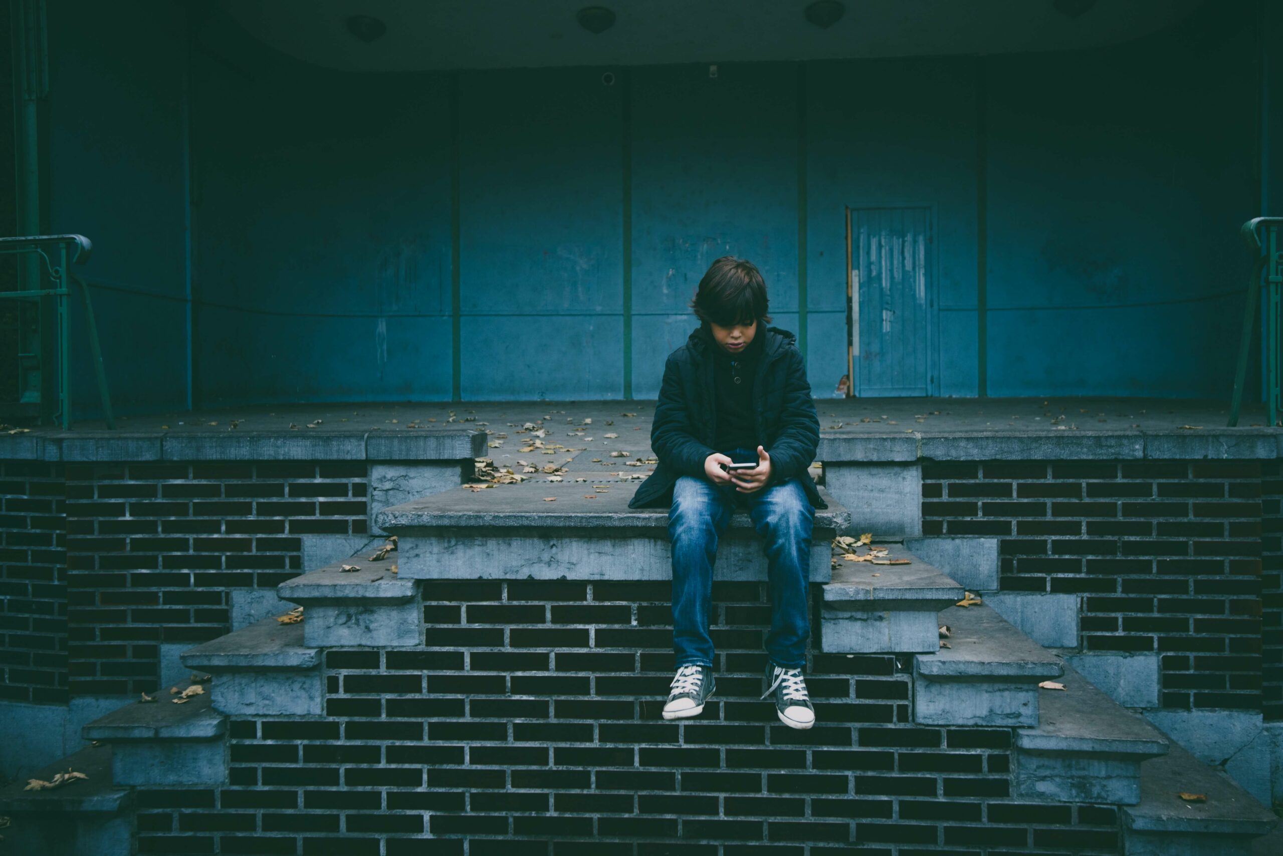 Young teenage boy sits on brick steps and looks down at a cellphone in his hand.