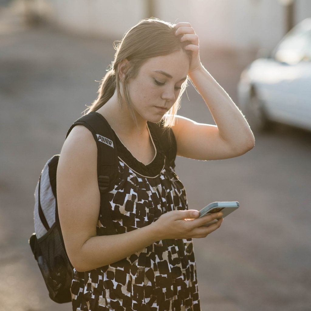 Photo of an american teenager, one hand on her head, the other holding a cell phone. Outside an american motel.