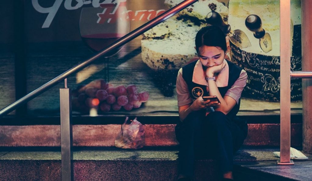 young Thai woman sitting outside using a smartphone, illustrating the prevalence of cybersex trafficking