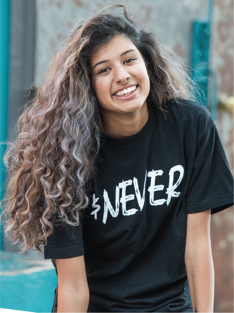 घुंघराले बालों वाली युवा अमेरिकी किशोरी एक्सोडस रोड टी शर्ट पहने हुए कैमरे पर मुस्कुराती है।