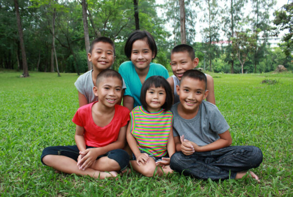 घास में बैठे थाई बच्चों का एक समूह मुस्कुरा रहा है, यह दिखा रहा है कि हम किस प्रकार के लोगों की सेवा करना चाहते हैं The Exodus Road थाईलैंड