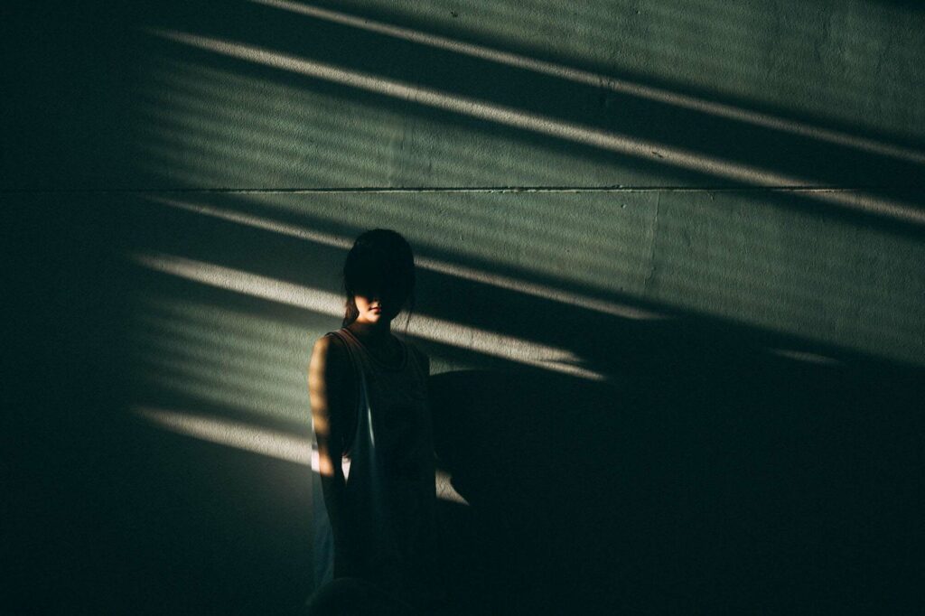 A female young adult stands against a concrete wall with shadows from a nearby window striking across her face.