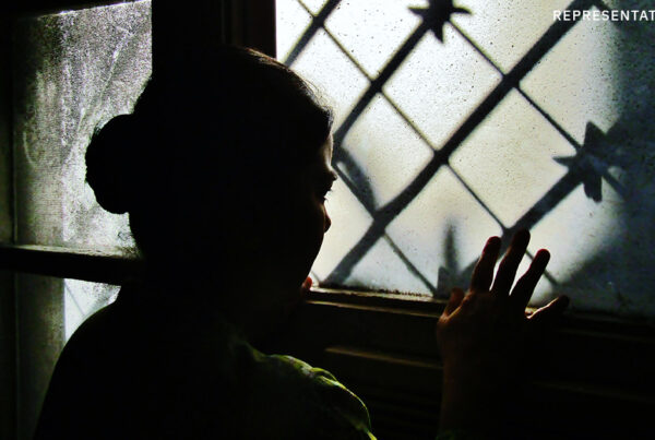 Indian girl looking out of a dark window, representing the confinement faced by the survivors from Operation 45 Minutes