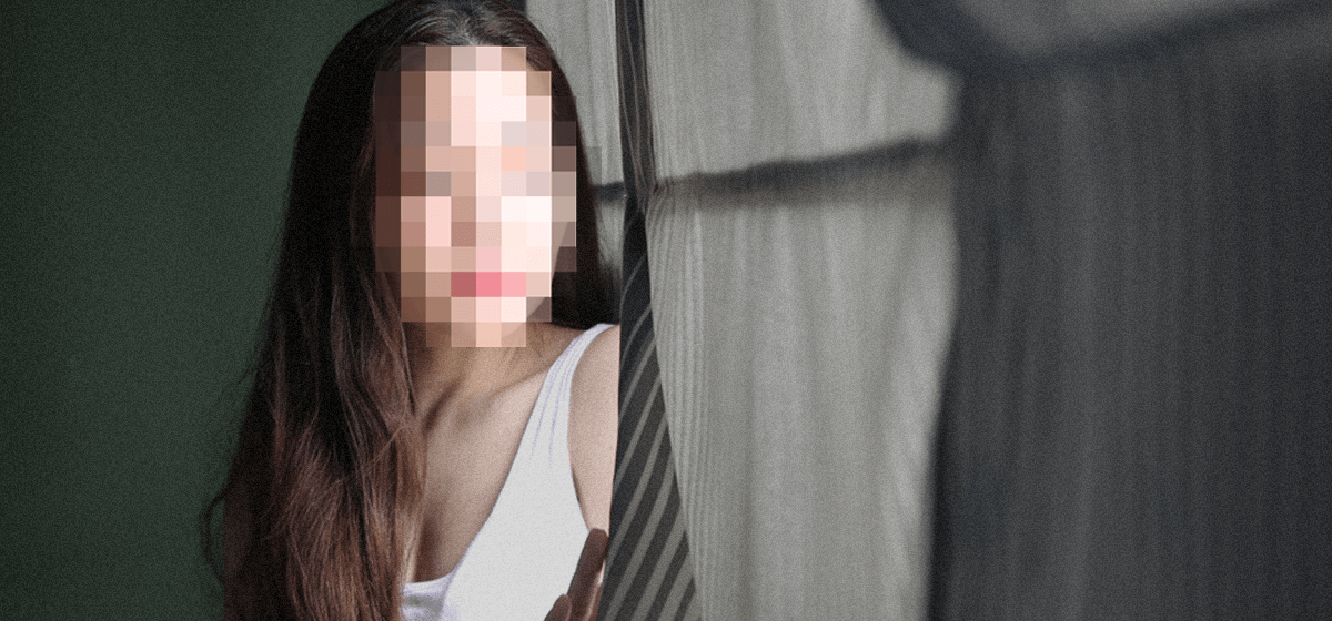 Young asian woman sits by window. Her face is pixelated, with a somber posture.