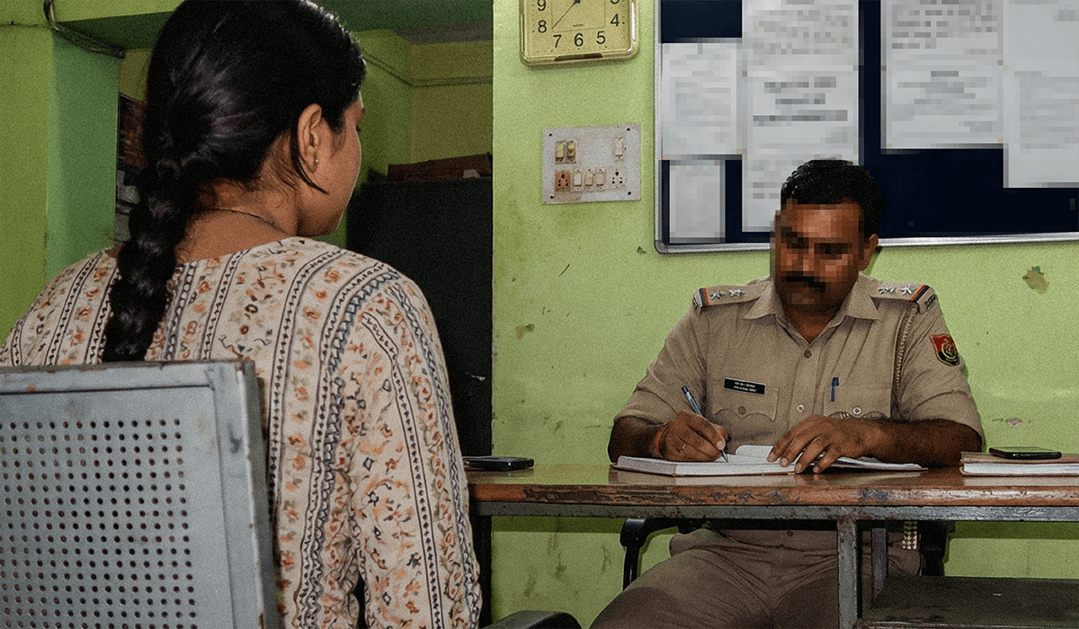 A young Indian girl sits in a police station, with her back to the camera. A police officer is writing notes in a notebook at a desk.