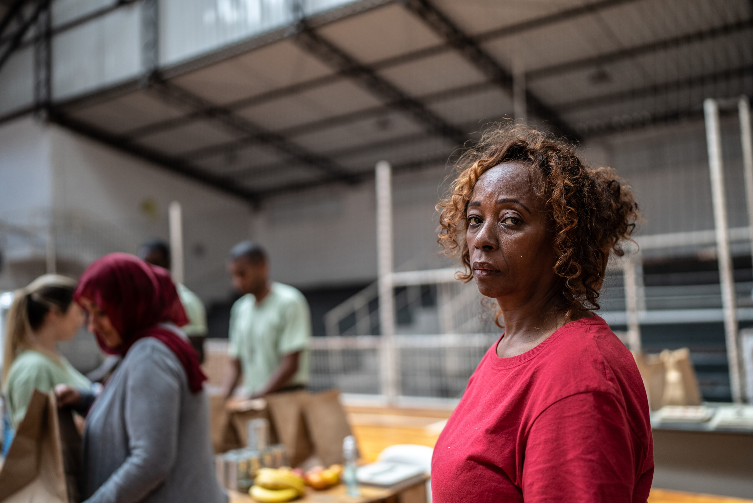 Woman in a red shirt looking at the camera while volunteers sort food donations in a community center