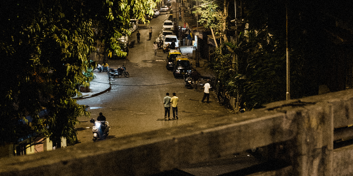 Two Indian men stand in the middle of a street lined by trees, at nighttime.