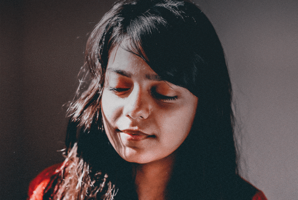 Teen girl from India looks down, sunlight hitting her face and lighting it up on a plain background.