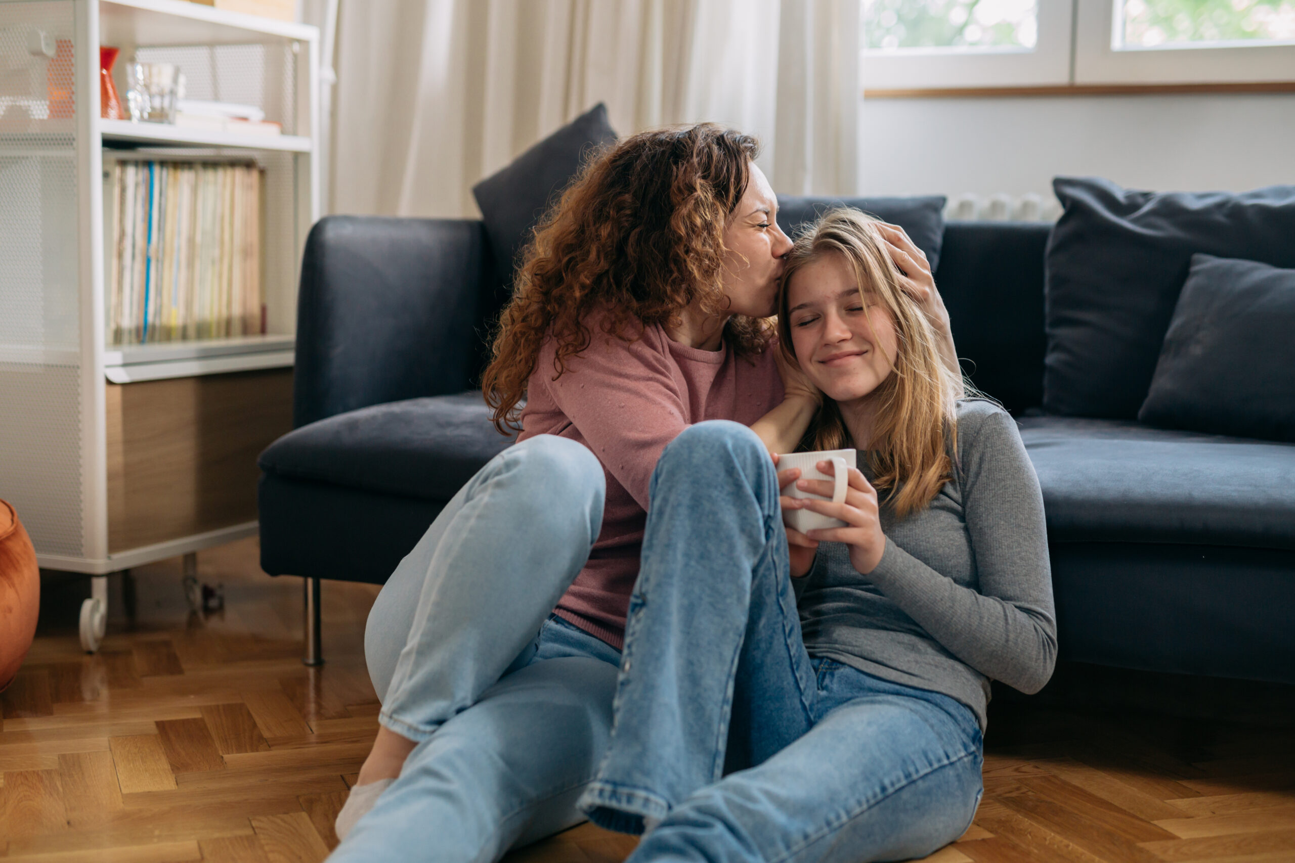 An adult tenderly embraces a teenager at home, both seated on the floor.