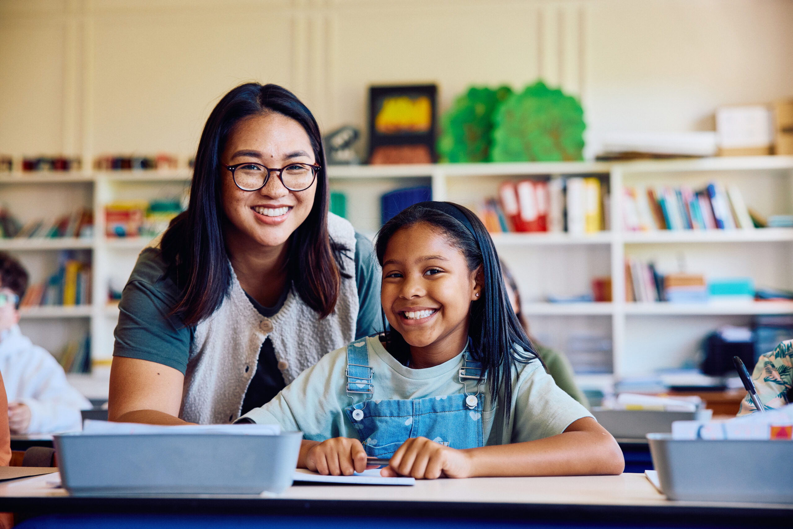 A teacher and student smile together at a classroom desk.