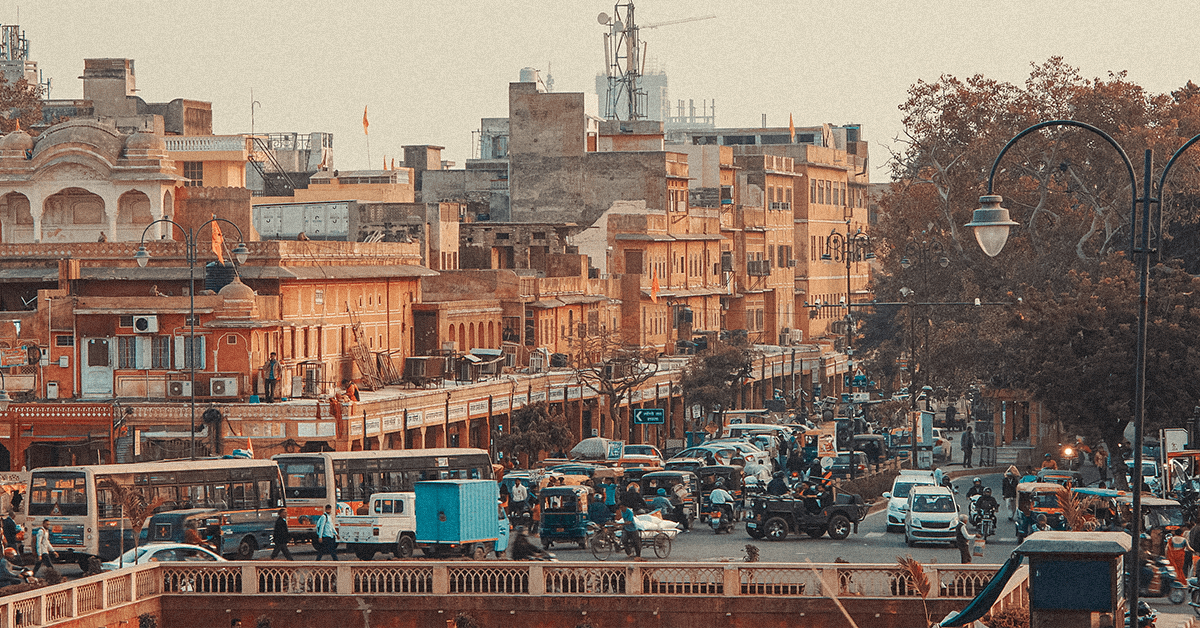 A vibrant street scene unfolds under the clear daylight in Jaipur, India. Ornate, historic buildings form the backdrop to the everyday bustle. Visible are numerous cars, motorcycles, and buses navigating the crowded roads. Many people walk along the sidewalks and crosswalks. The architecture showcases a mix of traditional designs and textures. The lighting conditions create a warm atmosphere enhancing the overall feel of the city. The scene could be useful in projects relating to travel, urban life, cityscapes, and transportation.