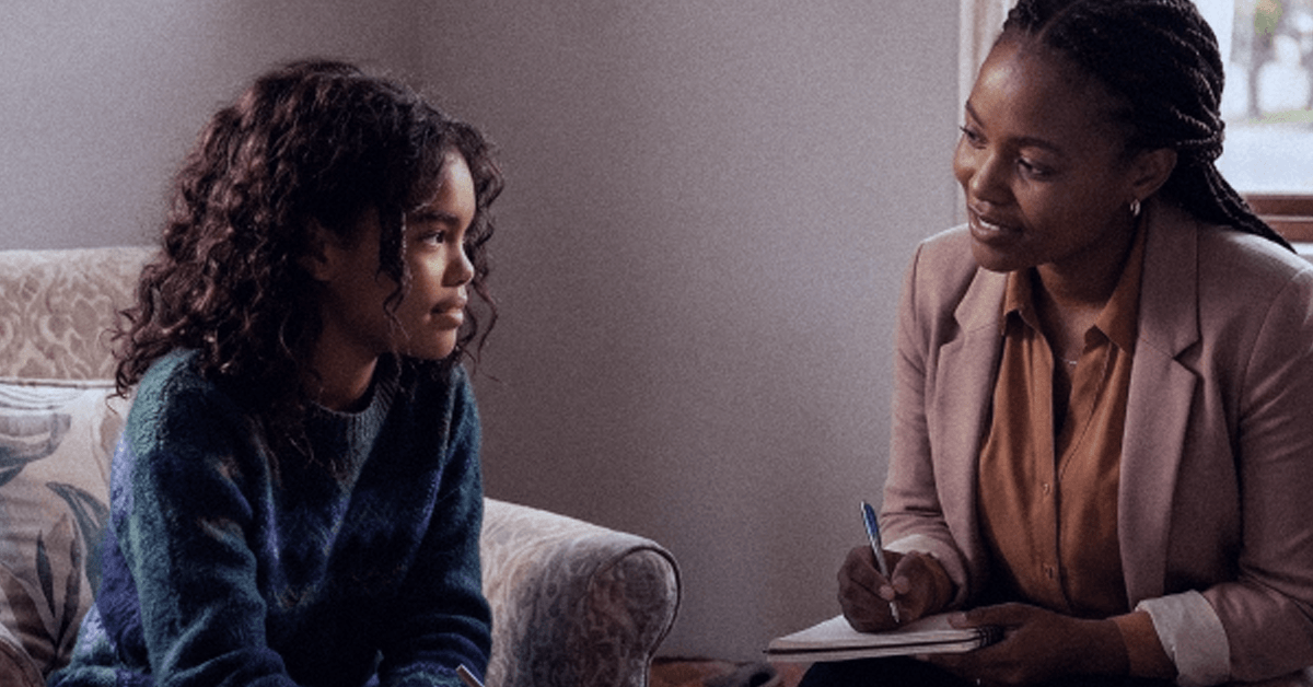 A 12-year-old girl sits with a therapist, who is smiling gently and taking notes.