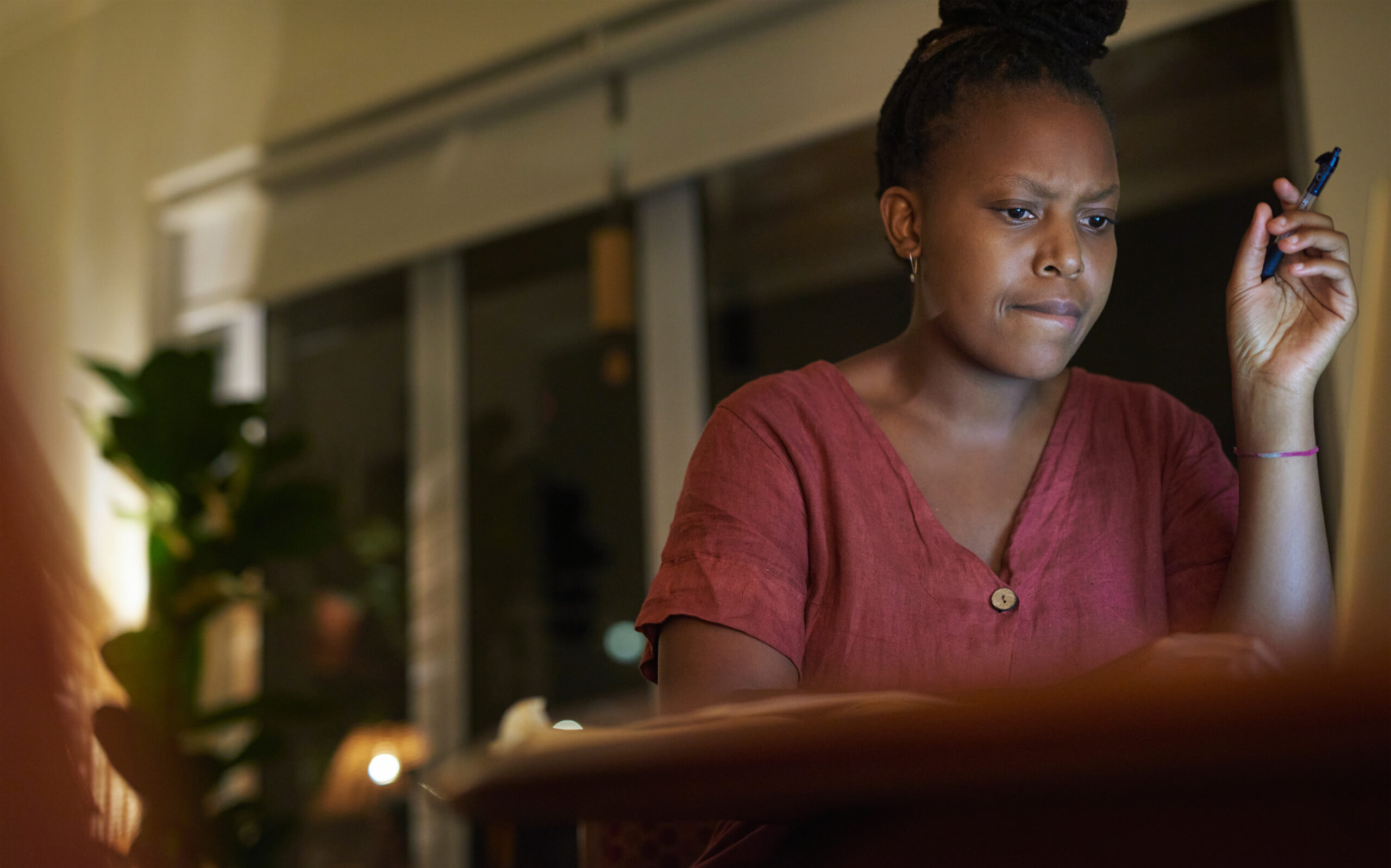 A woman sitting at a table in the evening, looking thoughtfully at a laptop screen with a pen in her hand