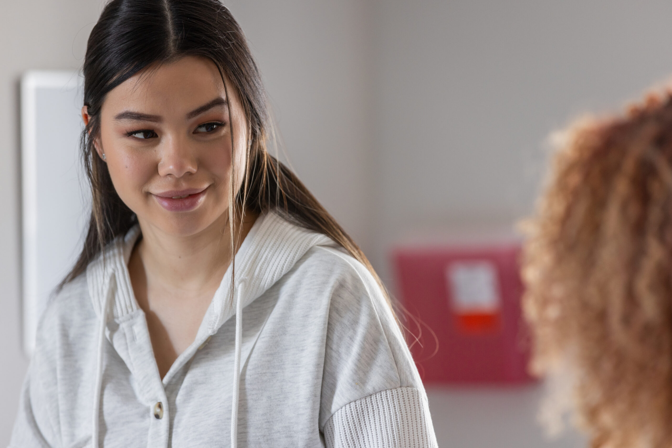 A young woman in a gray hoodie smiling gently as she listens to another person