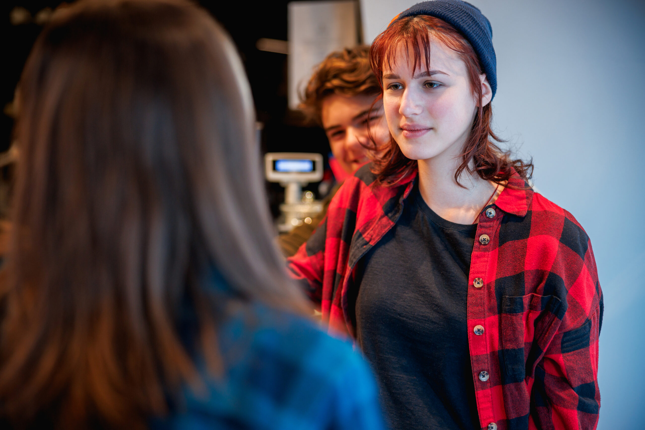 A young woman in a red plaid shirt and beanie listening to a friend, with another person standing behind her