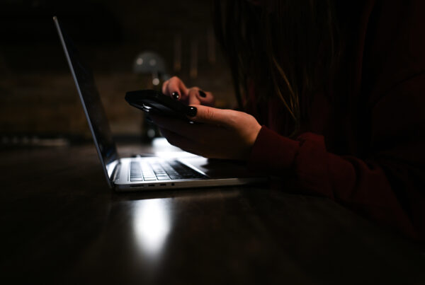 Hands in a dark room holding a phone over a laptop, representing a possible trafficking survivor